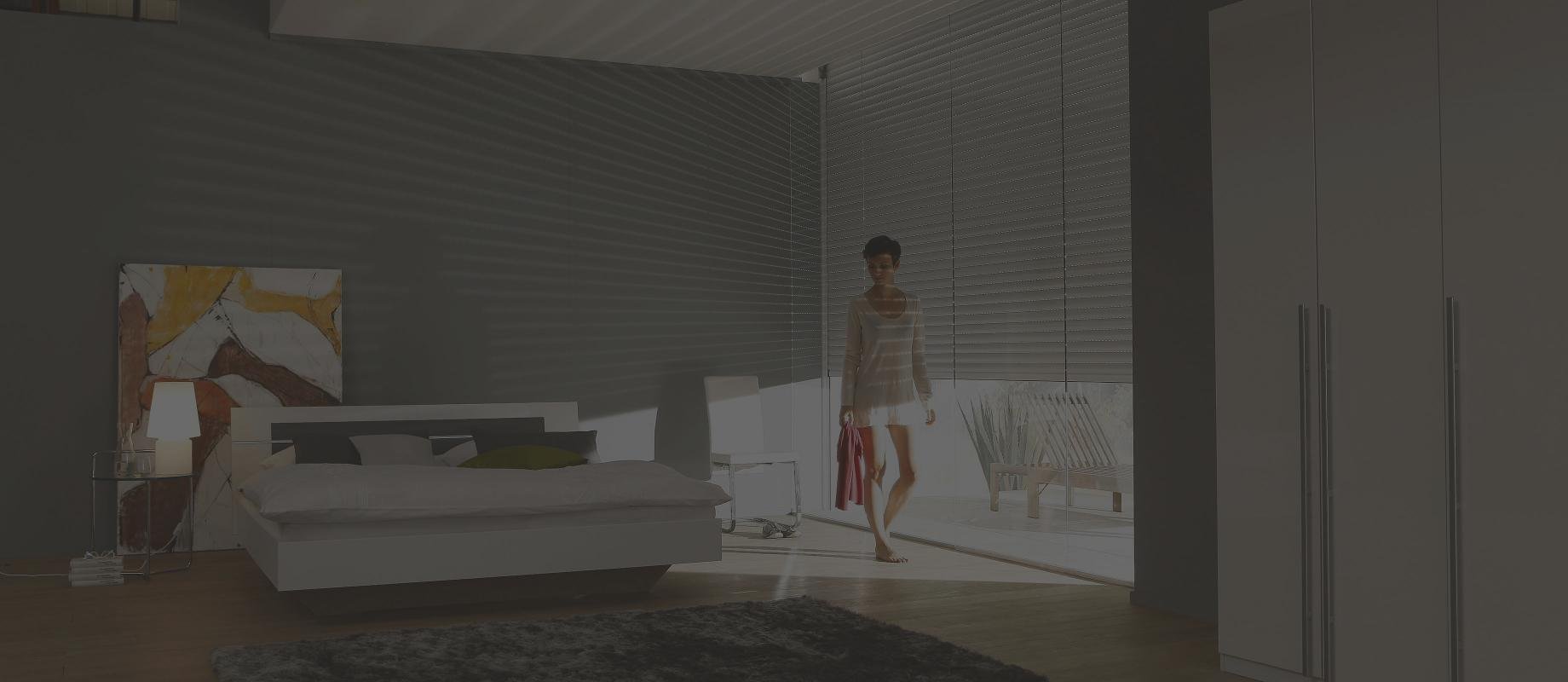 Woman stands in a beedroom in front of a window with half closed roller shutters