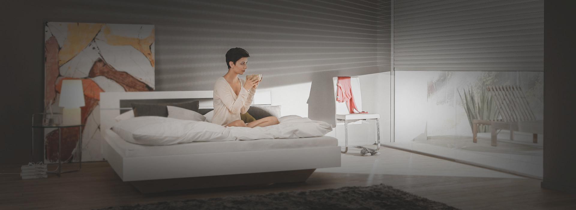 Woman sits on bed with half-closed roller shutters by daylight