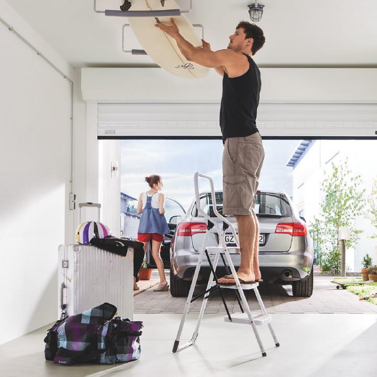 Optimal Use of Garage Space Man on ladder in open garage takes surfboard from the ceiling